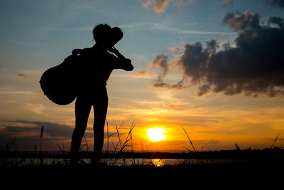 Silhouette woman carrying guitar case against sky during sunset