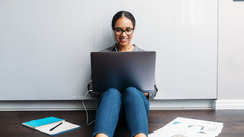 Smiling young woman using laptop while sitting on floor against wall