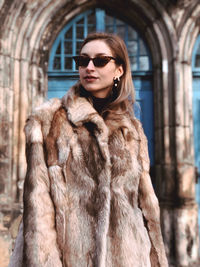 Fashionable young woman wearing fur coat and sunglasses while standing in city