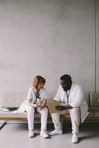 Full length of senior doctor discussing over clipboard with young male colleague sitting on sofa at hospital