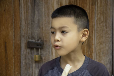 Boy looking away while eating food against wooden door