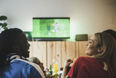 Smiling male and female watching soccer while sitting with friends at home