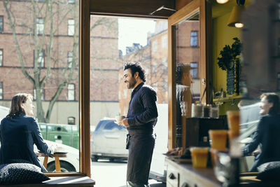 Smiling waiter talking with woman on sidewalk seen through glass at cafe