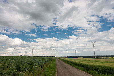 Street amidst wind turbines against cloudy sky