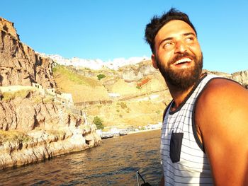 Cheerful young man looking away while standing by river against clear blue sky