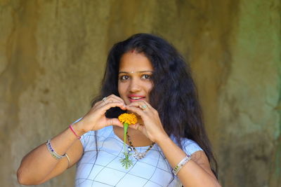A young woman holds a yellow marigold flower in her heart with fingers, two hands making heart sign