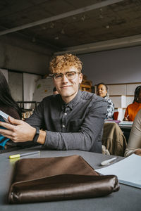 Portrait of smiling blond young male student sitting at desk in university classroom