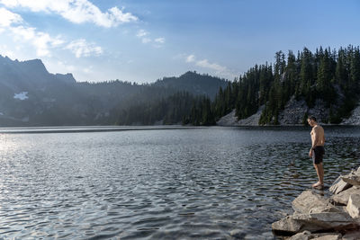 Side view of shirtless man standing by lake against sky