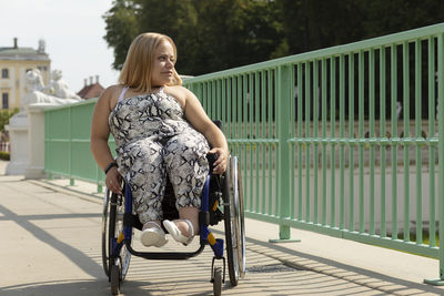 Beautiful young woman with short height walks over bridge in wheelchair in city at summer day