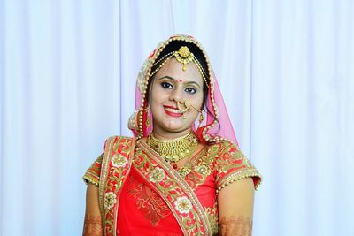 Portrait of smiling bride wearing jewelries and traditional clothing standing against curtain