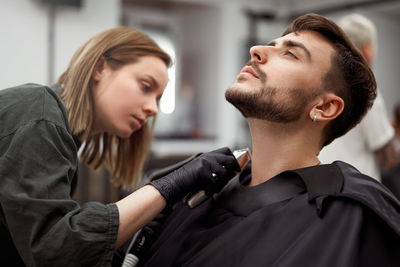 Portrait of young man who sitting in barbershop and woman hairdresser cut hair