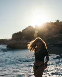 Young woman standing at beach against bright sun in clear sky