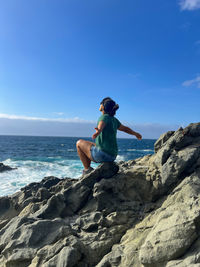 Rear view of woman sitting on rock at beach against blue sky