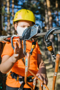 Focused girl in protective mask and helmet standing near tree in adventure park and putting carabine on safety rope during coronavirus