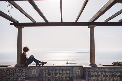 Side view of woman sitting by sea against sky