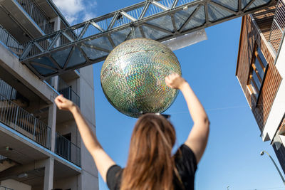 Low angle view of woman with arms raised against disco ball hanging from built structure