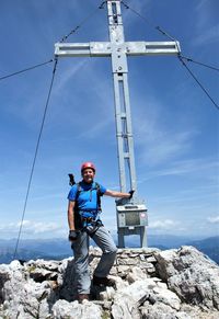 Male hiker standing by cross on cliff against blue sky