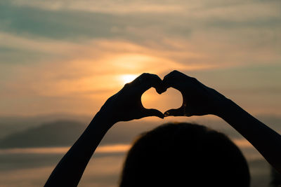 Close-up of hand making heart shape against sky during sunset