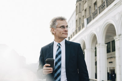 Confident businessman with disposable cup looking away while standing in city