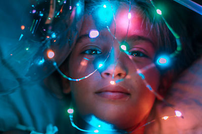 Close-up portrait of girl with illuminated string lights on bed