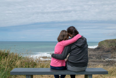 Rear view of mother and daughter sitting on bench at beach against sky