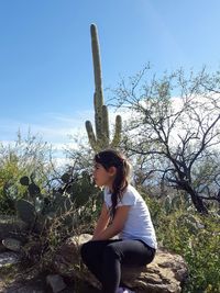 Side view of girl sitting on rock against blue sky