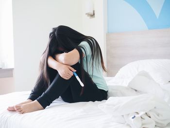 Worried woman with pregnancy test sitting on bed at home