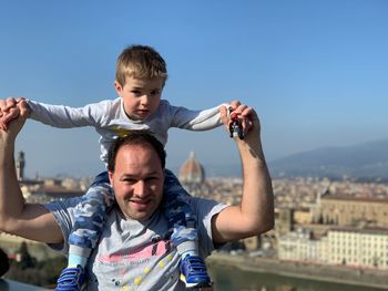 Portrait of smiling father carrying son on shoulders in city against sky