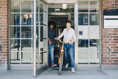 Full length of smiling businesswoman holding bicycle while walking with colleague through doorway at office