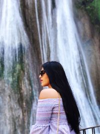 Side view of young woman with long hair standing against waterfall