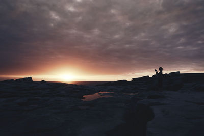 Silhouette woman on cliff by sea against sky during sunset