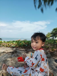 Portrait of young boy looking away while sitting on beach against sky