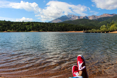 Low section of person relaxing on lakeshore against sky