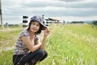 Portrait of young woman wearing cap crouching on grassy field against cloudy sky