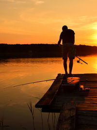 Full length of man holding fishing rod on pier by lake against sky during sunset