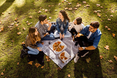 High angle view of happy friends eating while sitting on picnic blanket at park