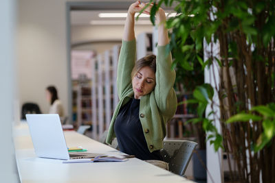 Tired female freelancer stretching arms relaxing while working on laptop in cozy coworking space