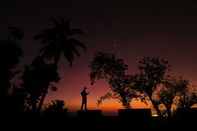 Silhouette man standing by palm tree against sky at sunset