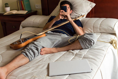 Bored man lying on a bed using phone after playing electric bass.