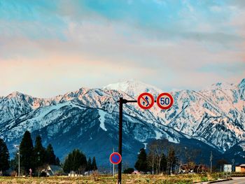 Road sign by snowcapped mountains against sky during winter