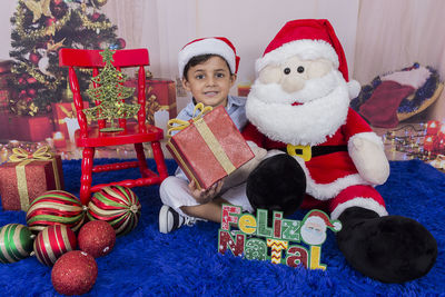 Portrait of smiling boy with santa claus toy and christmas presents sitting at home