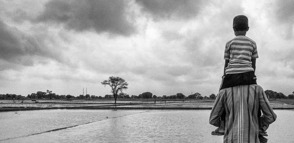 Rear view of father carrying son on shoulders while standing against lake