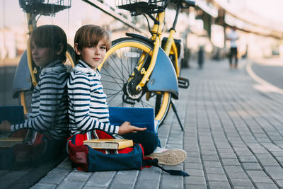 A cute schoolboy is sitting in the open air at school and holding a book in his hands. 