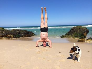 Shirtless man practicing headstand at beach against clear blue sky