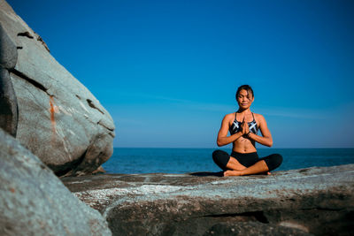 Young woman practicing yoga on rock at beach against blue sky