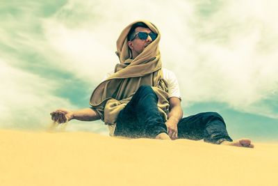 Low angle view of young man looking away while sitting on desert against sky