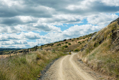 Dirt road amidst landscape against sky with a cyclist in the distance