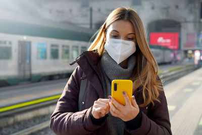 Girl in protective mask standing on the platform of train station with a phone buying ticket online