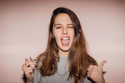 Portrait of beautiful young woman gesturing thumbs up while smoking cigarette against wall