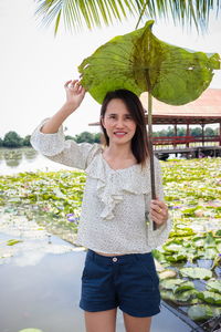 Portrait of smiling woman holding leaf while standing against plants in lake